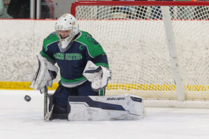 hockey puck sails towards goalie clad in green and blue.